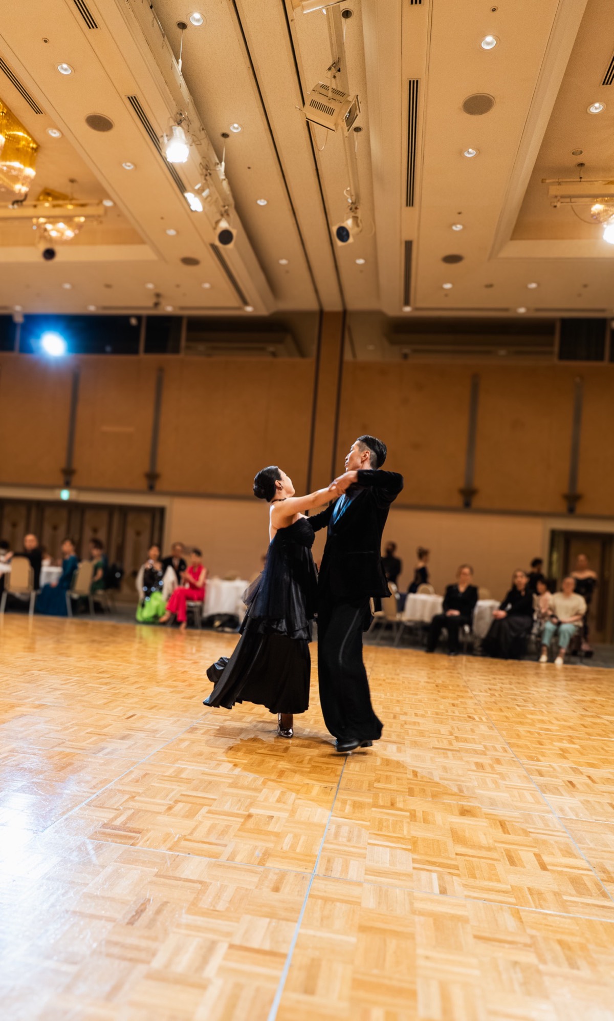 ホテルのボールルームでのパーティー。ひろいフロアで踊るひと組のカップルと、テーブルでくつろぐゲスト